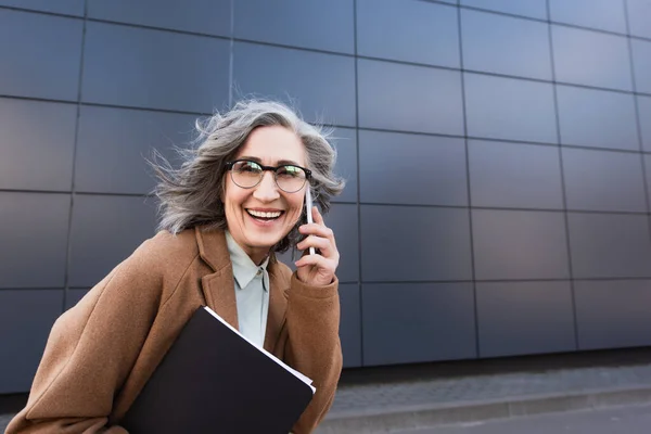 Middle aged businesswoman in coat talking on cellphone and holding documents while walking on urban street — Stock Photo