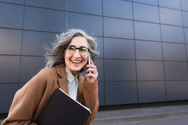 Femme d'affaires d'âge moyen en manteau parlant sur son téléphone portable et tenant des documents tout en marchant dans la rue urbaine — Photo de stock