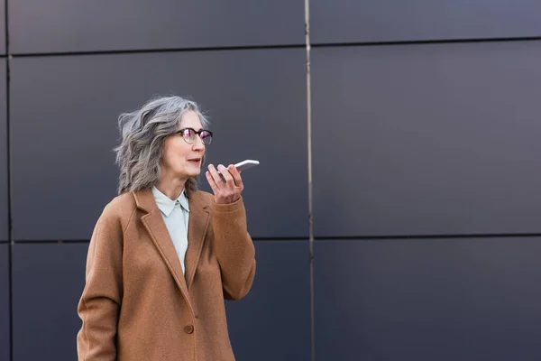 Reife Geschäftsfrau im Mantel nimmt Sprachnachricht auf Smartphone im Freien auf — Stockfoto