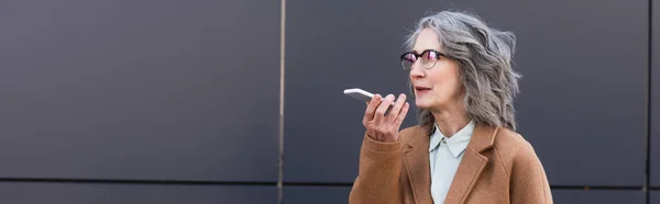 Grey haired businesswoman in coat recording voice message on smartphone near building outdoors, banner — Stock Photo