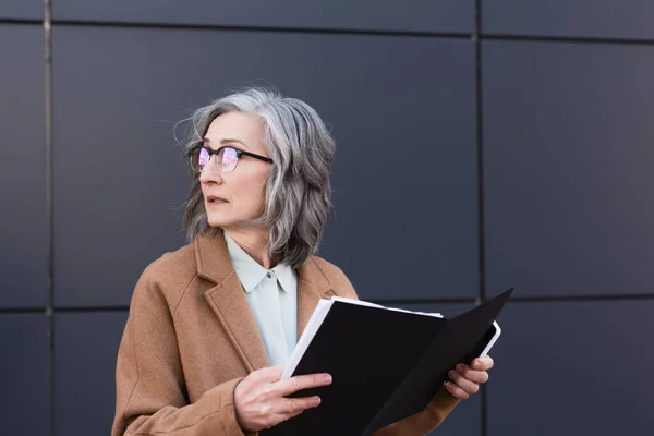 Mature businesswoman in coat holding paper folder and smartphone while looking away on urban street — Stock Photo