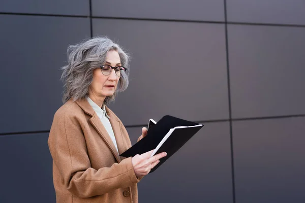 Femme d'affaires mature en lunettes et manteau tenant dossier en papier et téléphone portable à l'extérieur — Photo de stock