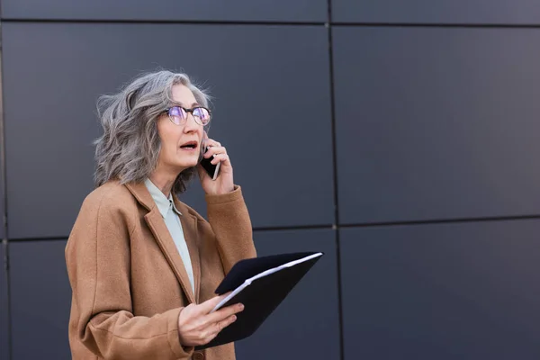 Gerente de pelo gris en abrigo que sostiene la carpeta de papel mientras habla en el teléfono inteligente en la calle urbana - foto de stock