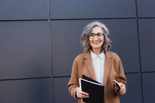 Mature businesswoman holding smartphone and paper folder while smiling at camera on urban street — Stock Photo