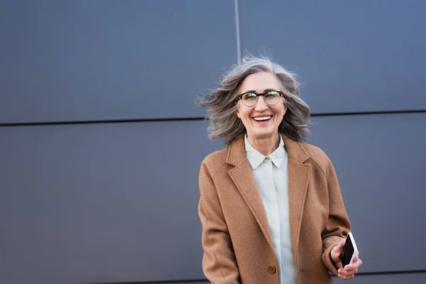 Зрелая деловая женщина в пальто держит смартфон с чистым экраном и улыбается на улице перед камерой — стоковое фото