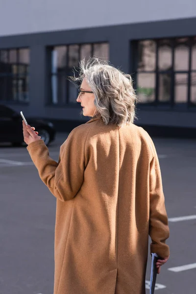 Side view of mature businesswoman in coat holding documents and using cellphone on urban street — Stock Photo