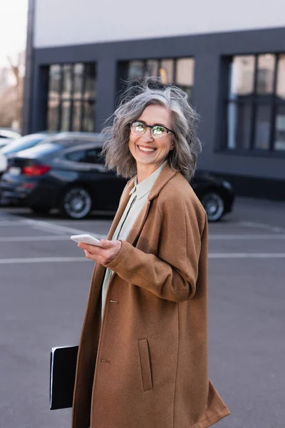 Mature businesswoman in coat holding mobile phone and paper folder on urban street — Stock Photo