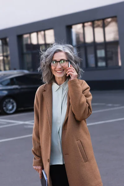 Cheerful mature businesswoman in coat talking on cellphone and holding documents on urban street — Stock Photo