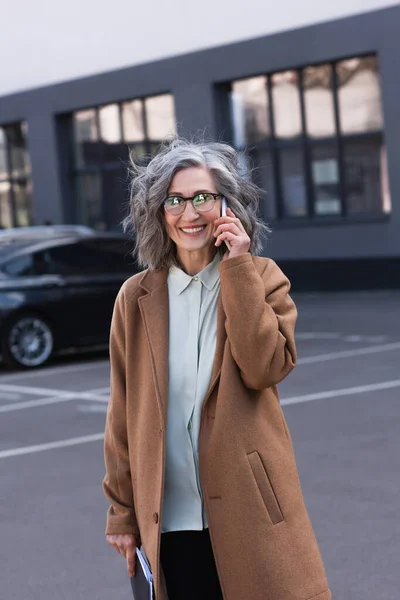 Cheerful mature businesswoman in coat talking on cellphone and holding documents on urban street — Stock Photo