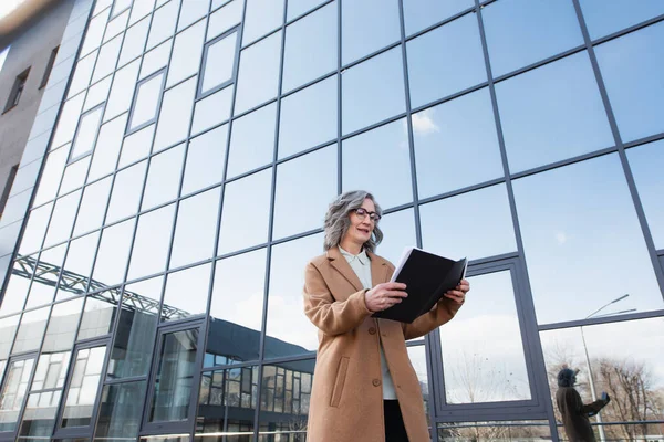 Vue en angle bas d'une femme d'affaires souriante en manteau regardant un dossier en papier et tenant un smartphone près d'un bâtiment à l'extérieur — Photo de stock