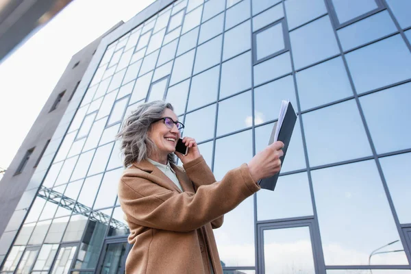 Tiefer Blickwinkel auf positive Geschäftsfrau, die auf dem Smartphone spricht und auf Papiermappe in der städtischen Straße blickt — Stockfoto