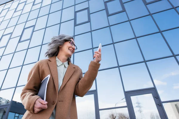 Visão de baixo ângulo de empresária alegre no casaco segurando pasta de papel e olhando para o celular na rua urbana — Fotografia de Stock