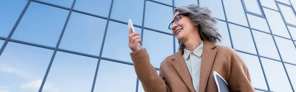 Vista de ángulo bajo de la mujer de negocios en abrigo sosteniendo papeles mientras mira el teléfono celular al aire libre, pancarta - foto de stock