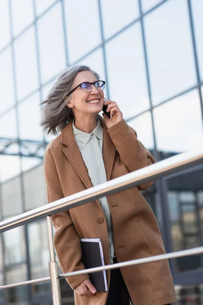 Cheerful businesswoman in coat holding documents and talking on mobile phone near railing outdoors — Stock Photo