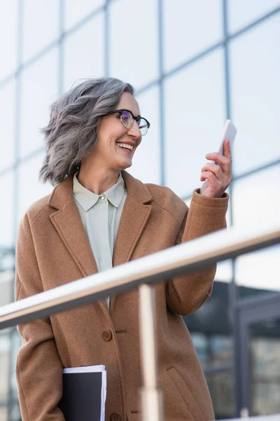 Happy businesswoman in coat holding documents and cellphone near blurred railing outdoors — Stock Photo