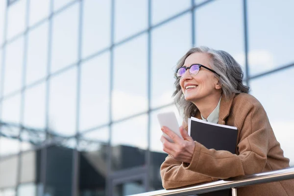 Зрелая деловая женщина в очках и пальто держит мобильный телефон и бумажную папку рядом с перилами на открытом воздухе — стоковое фото