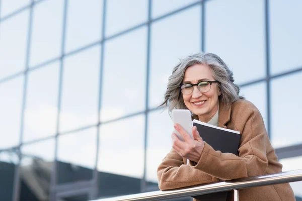 Grey haired manager in coat using smartphone while holding paper folder on urban street — Stock Photo