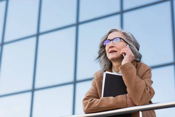 Middle aged businesswoman talking on cellphone and holding documents near railing on urban street — Stock Photo