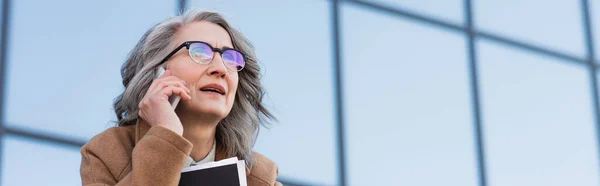 Empresária de cabelos grisalhos em óculos falando no celular e segurando pasta de papel ao ar livre, banner — Fotografia de Stock