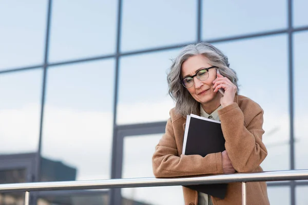 Upset businesswoman in coat talking on smartphone and holding papers on urban street — Stock Photo