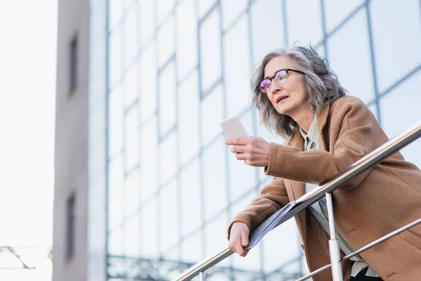 Businesswoman in coat holding papers and using smartphone near railing and building outdoors — Stock Photo