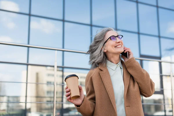 Feliz mujer de negocios senior hablando en el teléfono inteligente y sosteniendo la taza de papel en la calle urbana - foto de stock