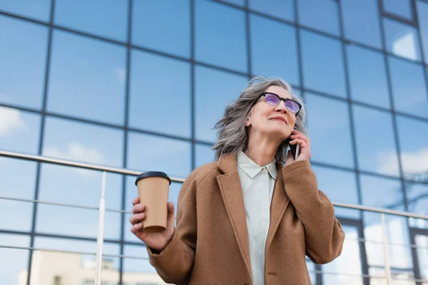 Зрелая деловая женщина в пальто и очках разговаривает по смартфону и держит кофе, чтобы выйти на улицу — стоковое фото