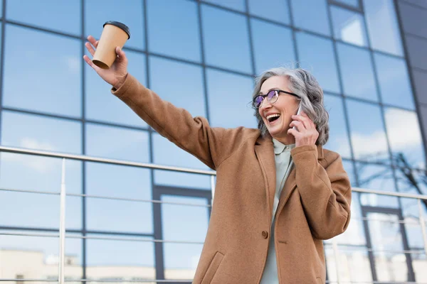 Счастливая седовласая деловая женщина в пальто разговаривает по мобильному телефону и держит выпивку на городской улице — стоковое фото