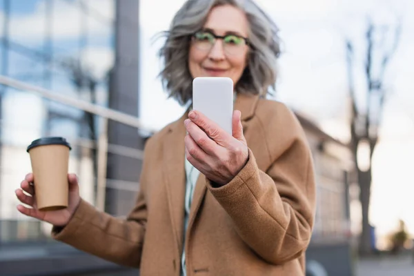 Smartphone in hand of blurred businesswoman in coat holding coffee to go outdoors — Stock Photo