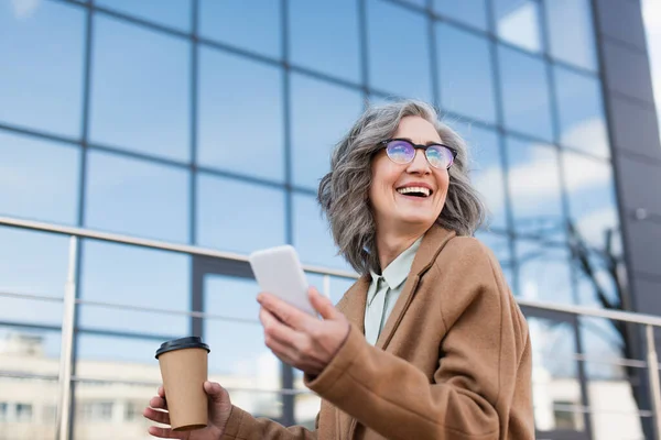 Joyful businesswoman in coat holding blurred smartphone and paper cup outdoors — Stock Photo
