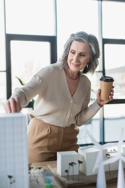 Mujer de negocios de pelo gris sonriente sosteniendo una taza de papel cerca de modelos borrosos de edificios en la oficina - foto de stock