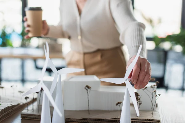 Vista recortada de empresaria borrosa con taza de papel tocando el modelo de turbina eólica en la oficina - foto de stock