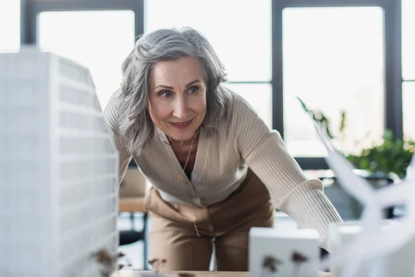 Sonriente mujer de negocios de mediana edad de pie cerca de modelos borrosas de edificios en la oficina - foto de stock