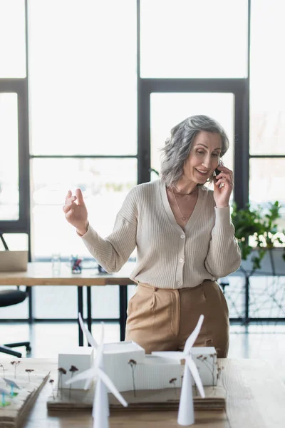 Весела зріла бізнес-леді розмовляє на смартфоні біля моделей будівель в офісі — стокове фото