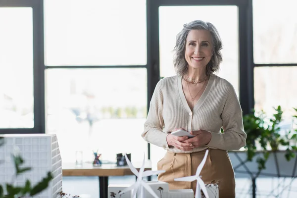 Femme d'affaires mature tenant smartphone et souriant à la caméra près des modèles de bâtiments dans le bureau — Photo de stock