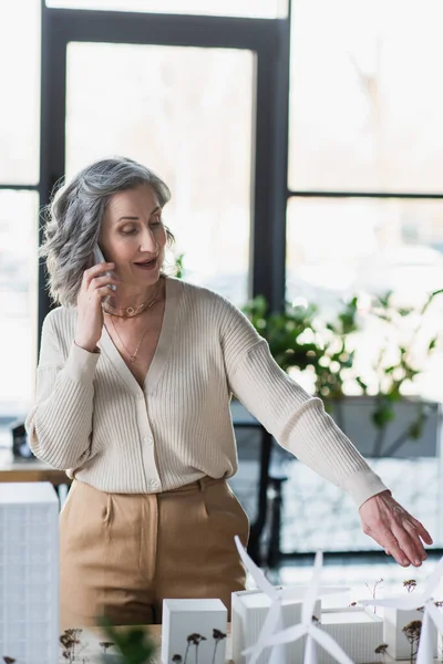 Empresaria hablando en smartphone y apuntando con la mano a los modelos de edificios en la oficina - foto de stock