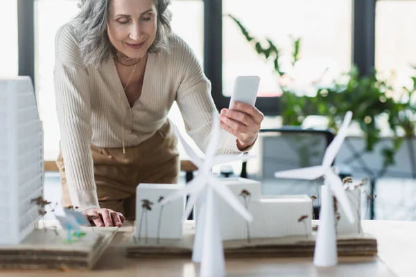 Smiling businesswoman holding cellphone near models of buildings and wind turbines in office — Stock Photo