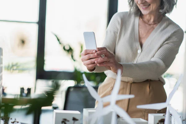 Vista recortada de una mujer de negocios madura usando un teléfono móvil cerca de modelos borrosos de turbinas eólicas en la oficina - foto de stock