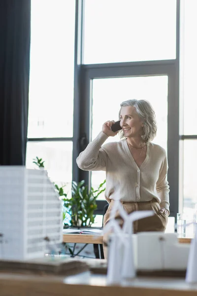 Femme d'affaires positive parlant sur un téléphone portable près de modèles flous de bâtiments dans le bureau — Photo de stock