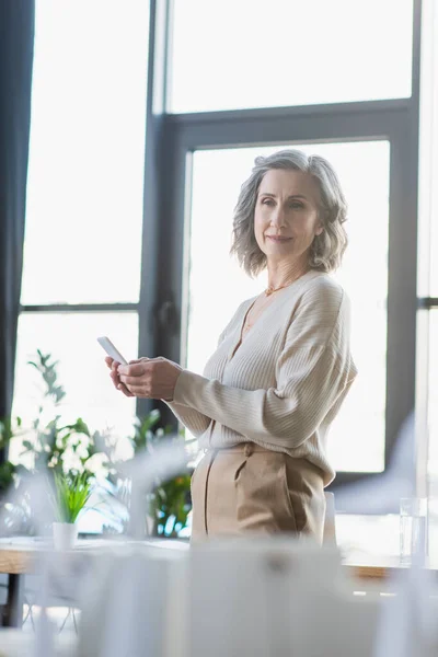 Femme d'affaires aux cheveux gris utilisant un téléphone portable près de modèles flous de bâtiment dans le bureau — Photo de stock