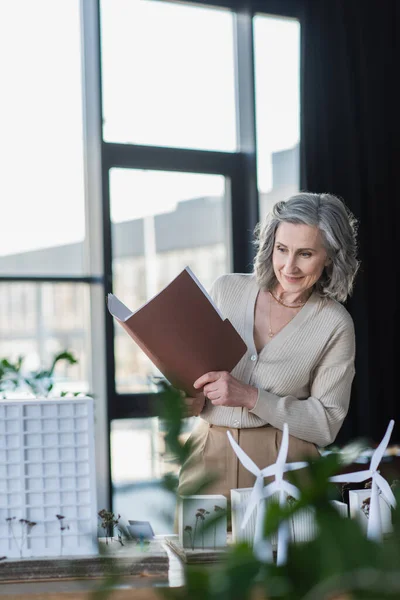 Femme d'affaires mature tenant dossier papier près des modèles de bâtiment dans le bureau — Photo de stock