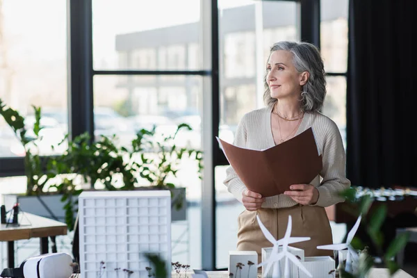 Mature businesswoman holding paper folder near vr headset and models of buildings in office — Stock Photo