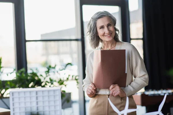 Empresária de cabelos grisalhos segurando pasta de papel e sorrindo para a câmera perto de modelos de edifícios no escritório — Fotografia de Stock