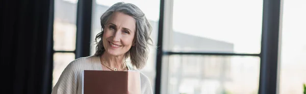 Middle aged businesswoman holding paper folder and smiling at camera in office, banner — Stock Photo