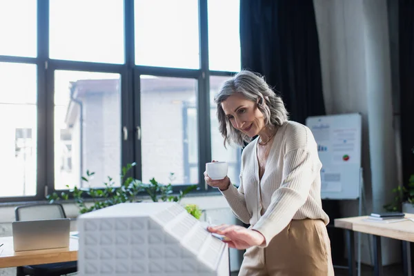 Enthousiaste femme d'affaires mature tenant tasse près du modèle de bâtiment dans le bureau — Photo de stock