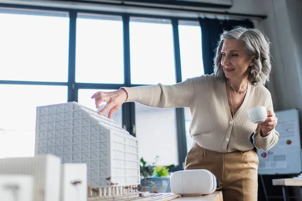 Mulher de negócios madura segurando xícara de café e gestos perto de modelos de edifícios e fones de ouvido vr no escritório — Fotografia de Stock