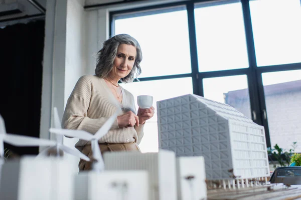 Бізнес-леді тримає чашку кави біля розмитих моделей будівель в офісі — стокове фото
