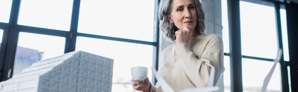 Nachdenkliche Geschäftsfrau hält Tasse in der Nähe Modell des Gebäudes im Büro, Banner — Stockfoto