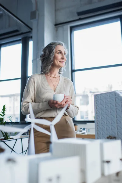 Happy mature businesswoman holding cup near blurred models of buildings in office — Stock Photo