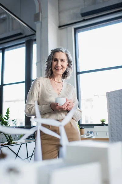 Positive businesswoman with coffee cup looking at camera near blurred models of buildings in office — Stock Photo